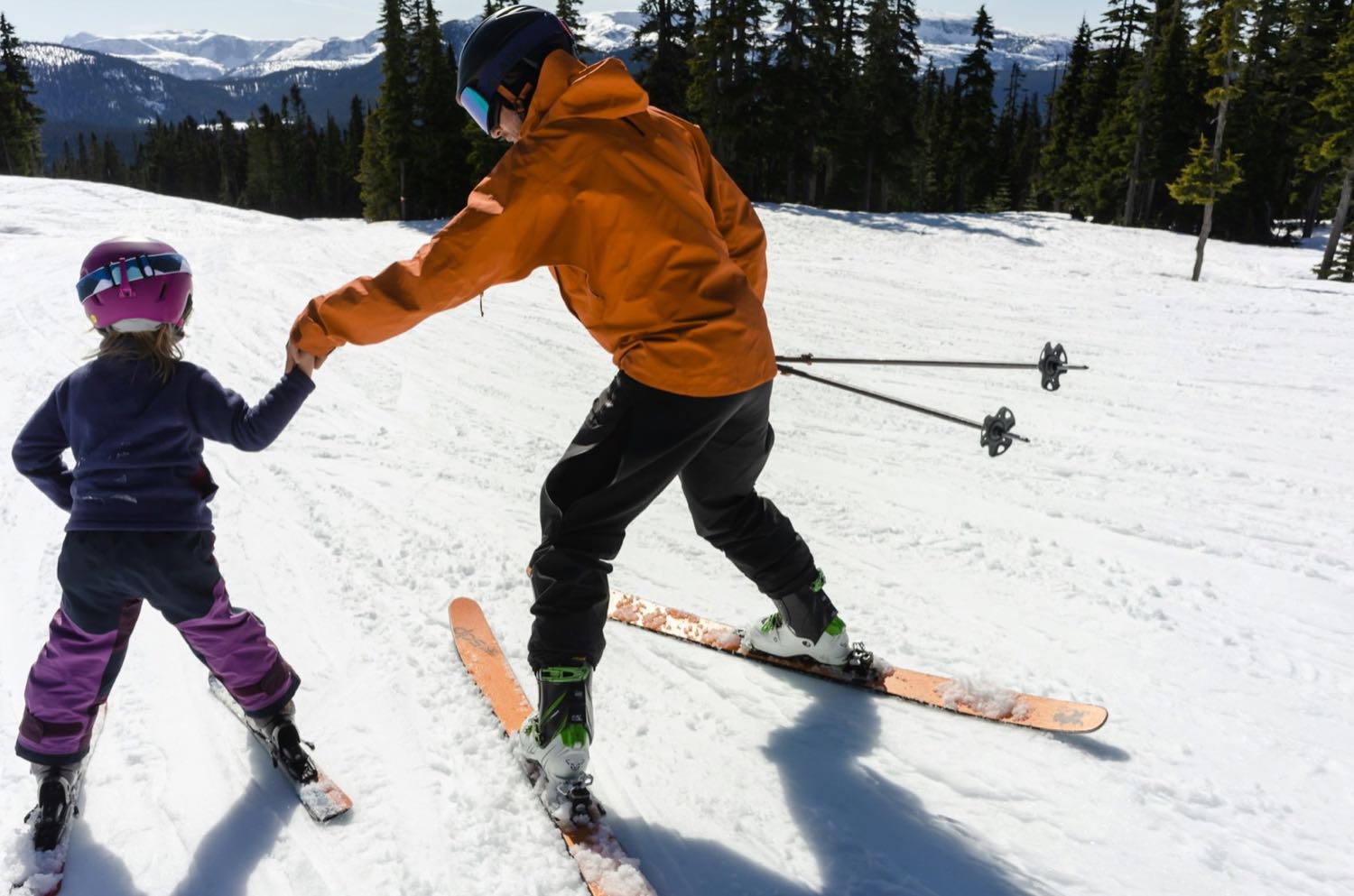 An adult holding a small child's hand as she takes her first turns on a gentle snowy slope