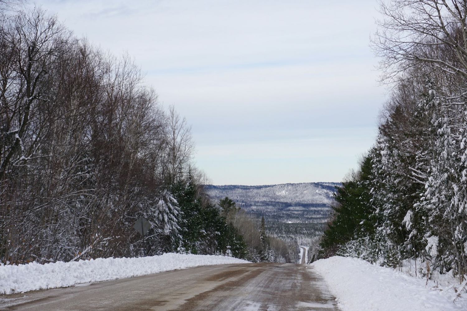A snow-banked two-lane road descending through winter forest towards forested hills
